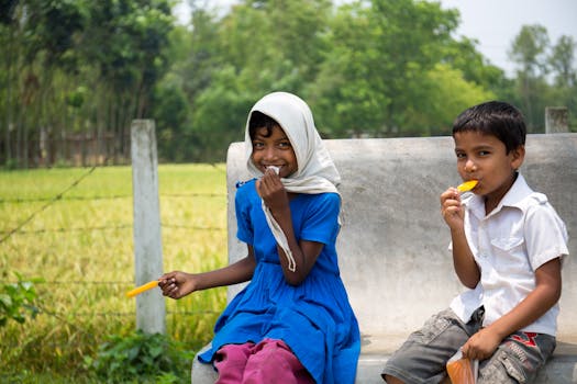 Two children enjoy ice pops while sitting on a bench outdoors, smiling happily in the sun.